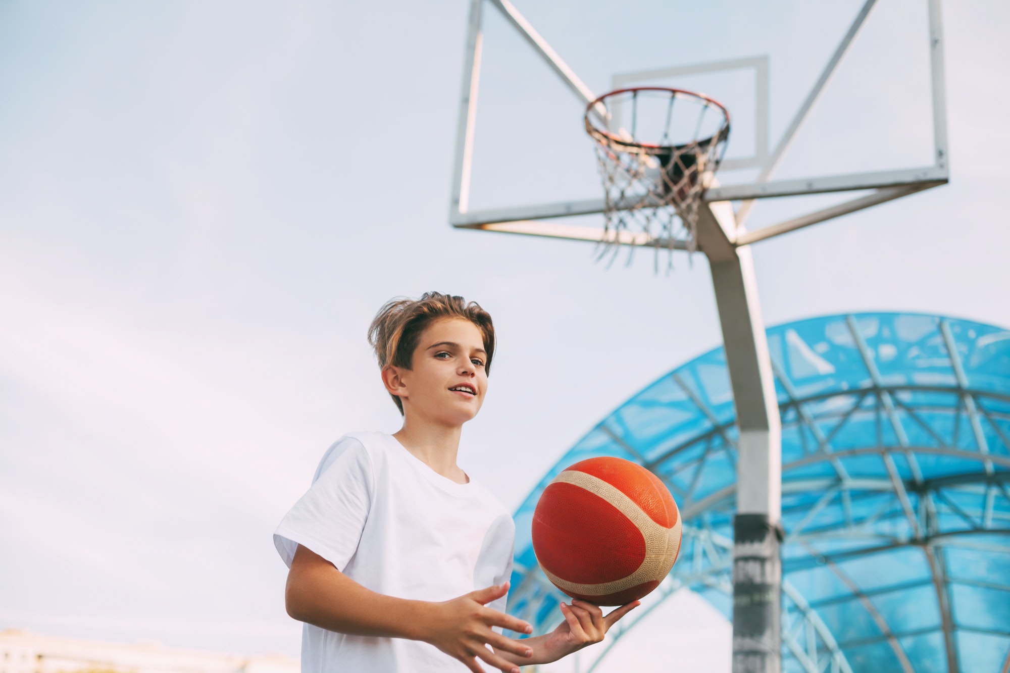A young teenage basketball player in a white t-shirt stands on the basketball court